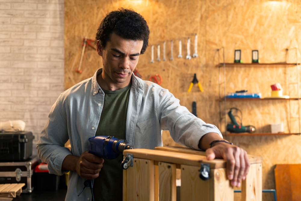 Man using a power drill to assemble a wooden crate in a workshop with tools hanging on the wall behind him.