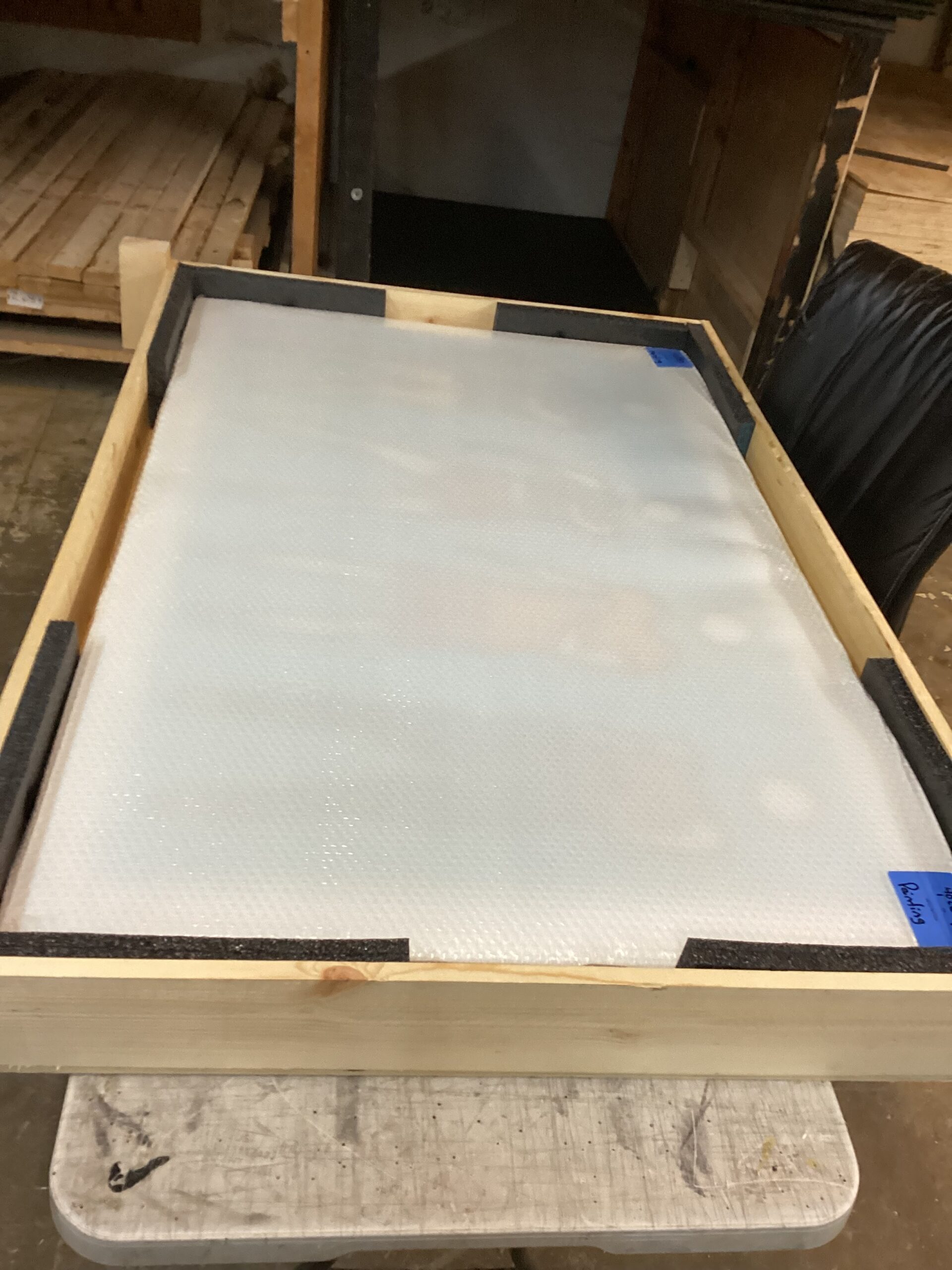 A large rectangular panel wrapped in bubble wrap is placed inside a shallow wooden crate lined with black foam padding. The crate rests on a worktable in a workshop, with stacked wood and tools visible in the background.