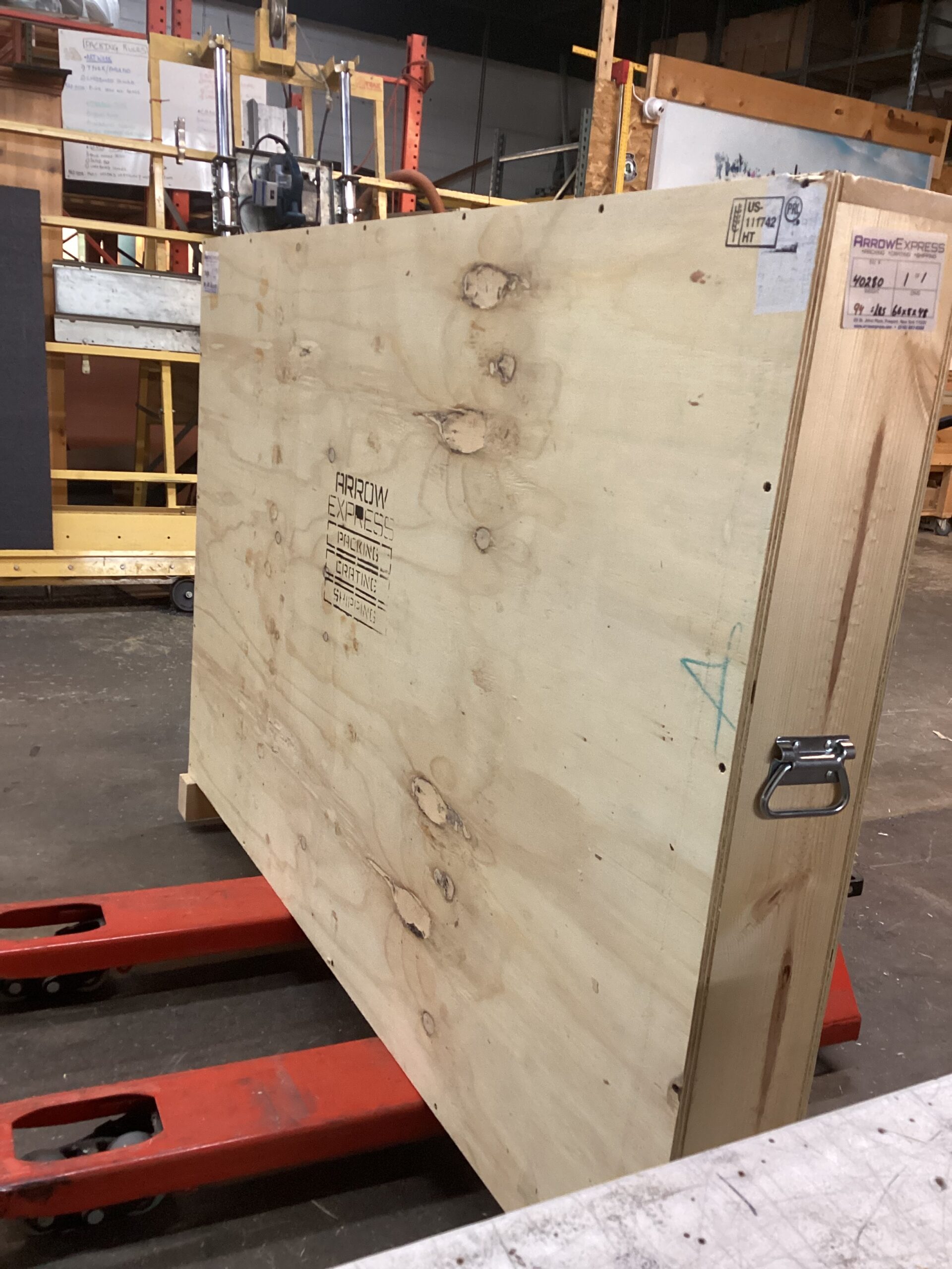 A large plywood shipping crate labeled “Arrow Express” stands upright on the red forks of a pallet jack in a workshop. The crate has metal side handles and shipping labels attached, with shelving, equipment, and industrial materials visible in the background.