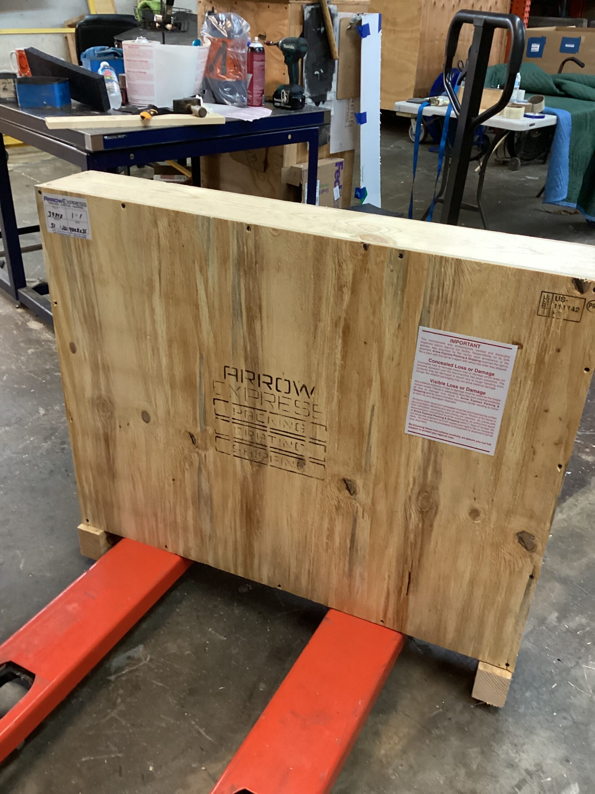 A sealed wooden shipping crate labeled “Arrow Express” sits on the red forks of a pallet jack in a workshop. The crate has warning and shipping notices attached, with tools, tables, and packing materials visible in the background.
