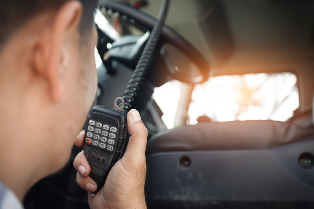 Close-up of a person inside a vehicle holding and speaking into a handheld radio microphone, with sunlight visible through the windshield.