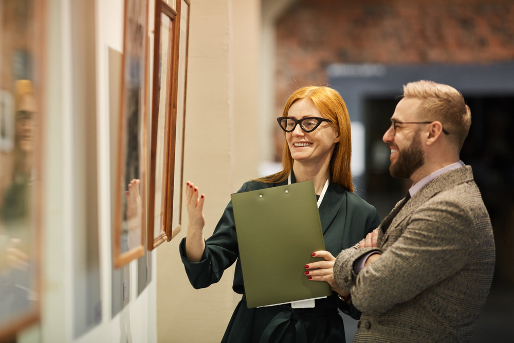 Two people stand in an art gallery, smiling and discussing framed artwork on the wall. One person holds a clipboard and gestures toward a piece while the other listens, with softly lit gallery walls and additional artworks in the background.