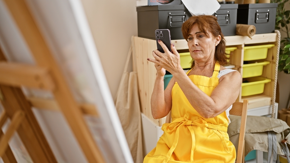 A woman wearing a yellow apron sits in an art studio, holding up a smartphone to photograph or examine a canvas on an easel. Shelving with storage boxes and art supplies is visible in the background.