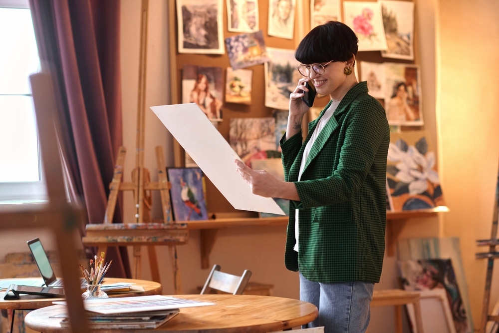 A person wearing glasses and a green jacket stands in an art studio, smiling while talking on a phone and holding a large sheet of paper. Paintings and sketches cover the wall behind them, with an easel, table, and art supplies visible nearby.