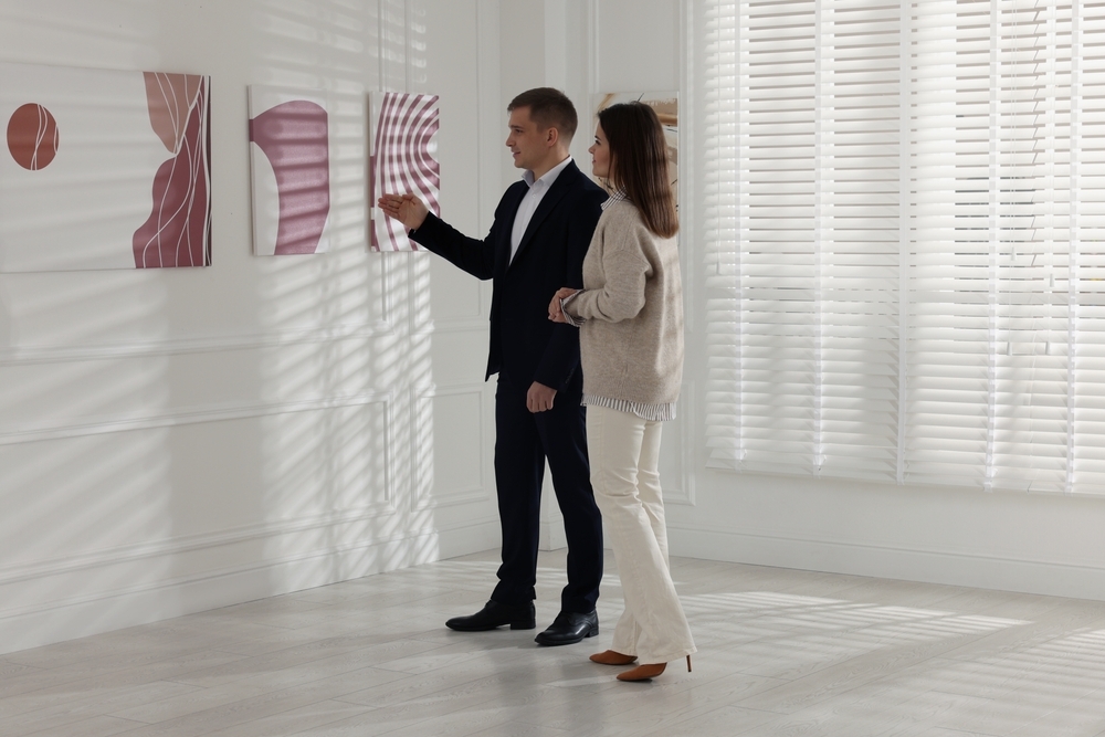 A man and woman stand in a bright, minimalist room with white walls and window blinds, looking at three abstract pink art pieces displayed on the wall. The man gestures toward one of the artworks while the woman stands beside him, observing.