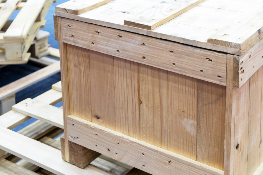 A sturdy wooden shipping crate made of slatted boards and secured with nails sits on a pallet, with additional wooden pallets and crates visible in the background.